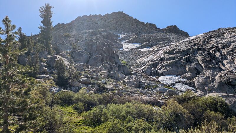 The image shows a rocky mountain landscape under a bright, sunny sky. Patches of snow are visible on the mountain's slopes, interspersed with areas of exposed rock and sparse vegetation. In the foreground, there are green bushes and a few trees. The overall impression is one of a rugged, natural environment.